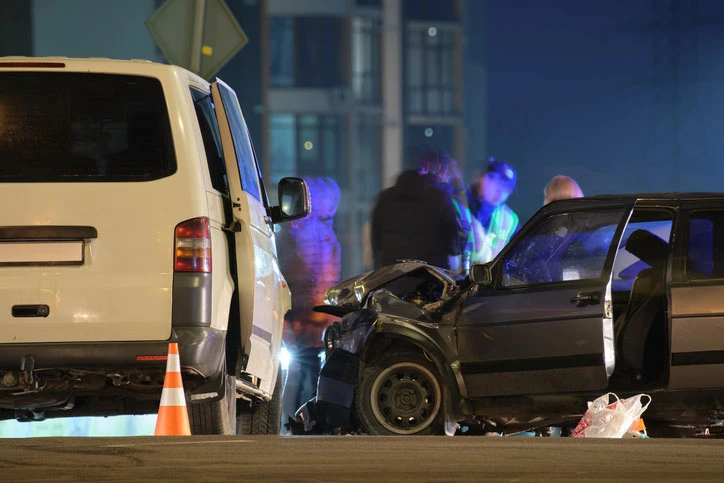 Nighttime crash scene showing a damaged sedan after colliding with a white van, with emergency responders and police lights in the background.